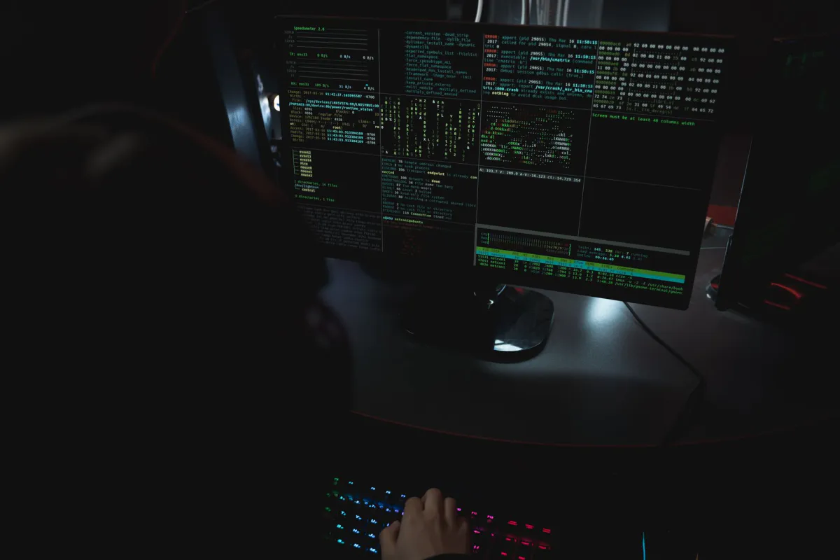 Écrans d'ordinateur affichant des lignes de code et des terminaux dans une pièce sombre, avec un clavier rétroéclairé au premier plan