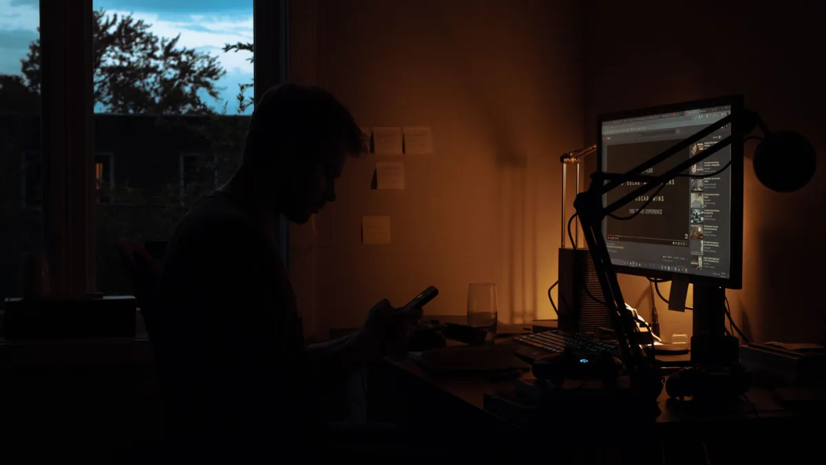 Silhouette d'une personne assise à un bureau le soir, consultant son téléphone face à un écran d'ordinateur allumé dans une pièce peu éclairée