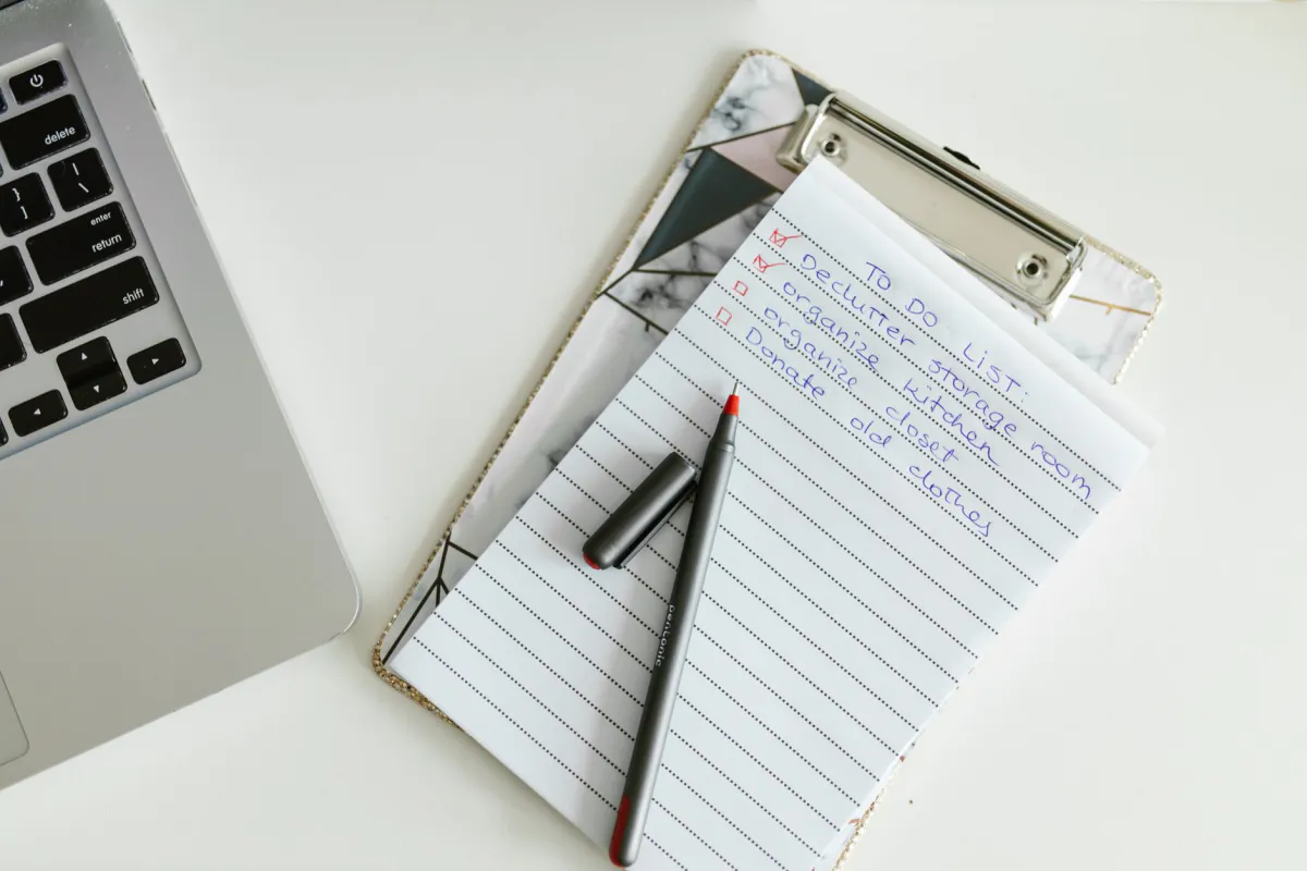 Illustration d'une liste de tâches manuscrite sur un porte-bloc, avec un stylo posé dessus et un ordinateur portable à côté