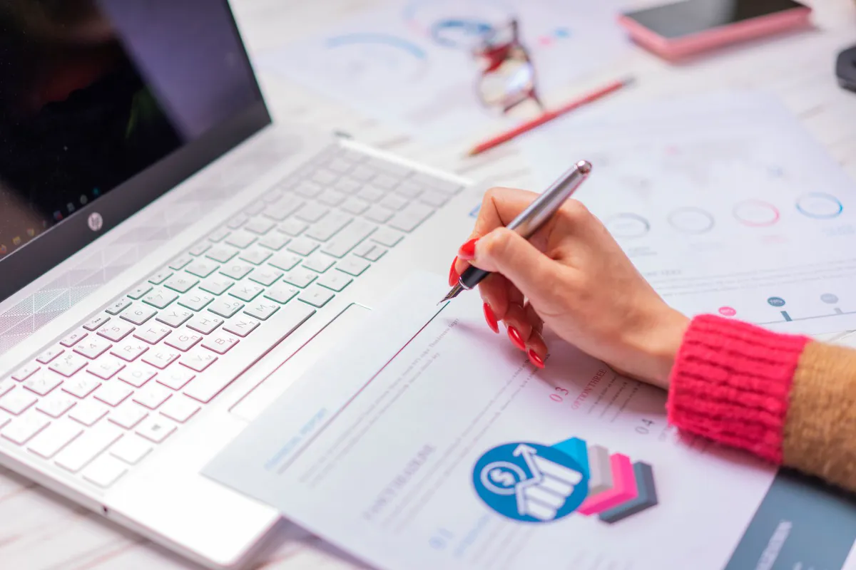 Main tenant un stylo au-dessus de graphiques de coûts imprimés, à côté d'un ordinateur portable