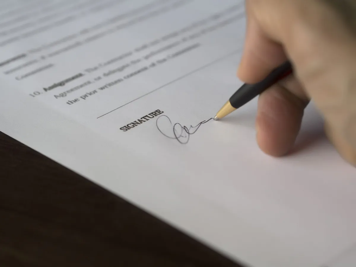 Illustration of a hand signing a contract document with a pen, signature line visible on the page
