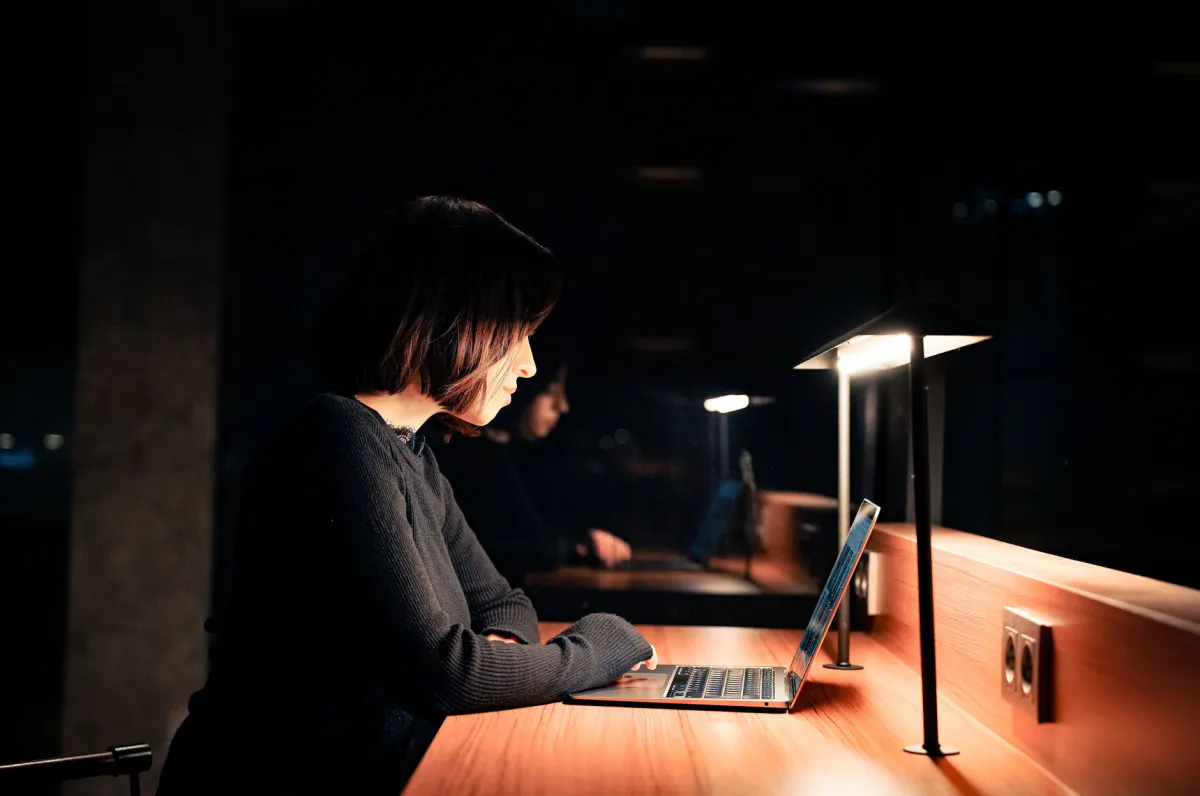 Person working on a laptop late at night, lit by a desk lamp in a dark room, illustration