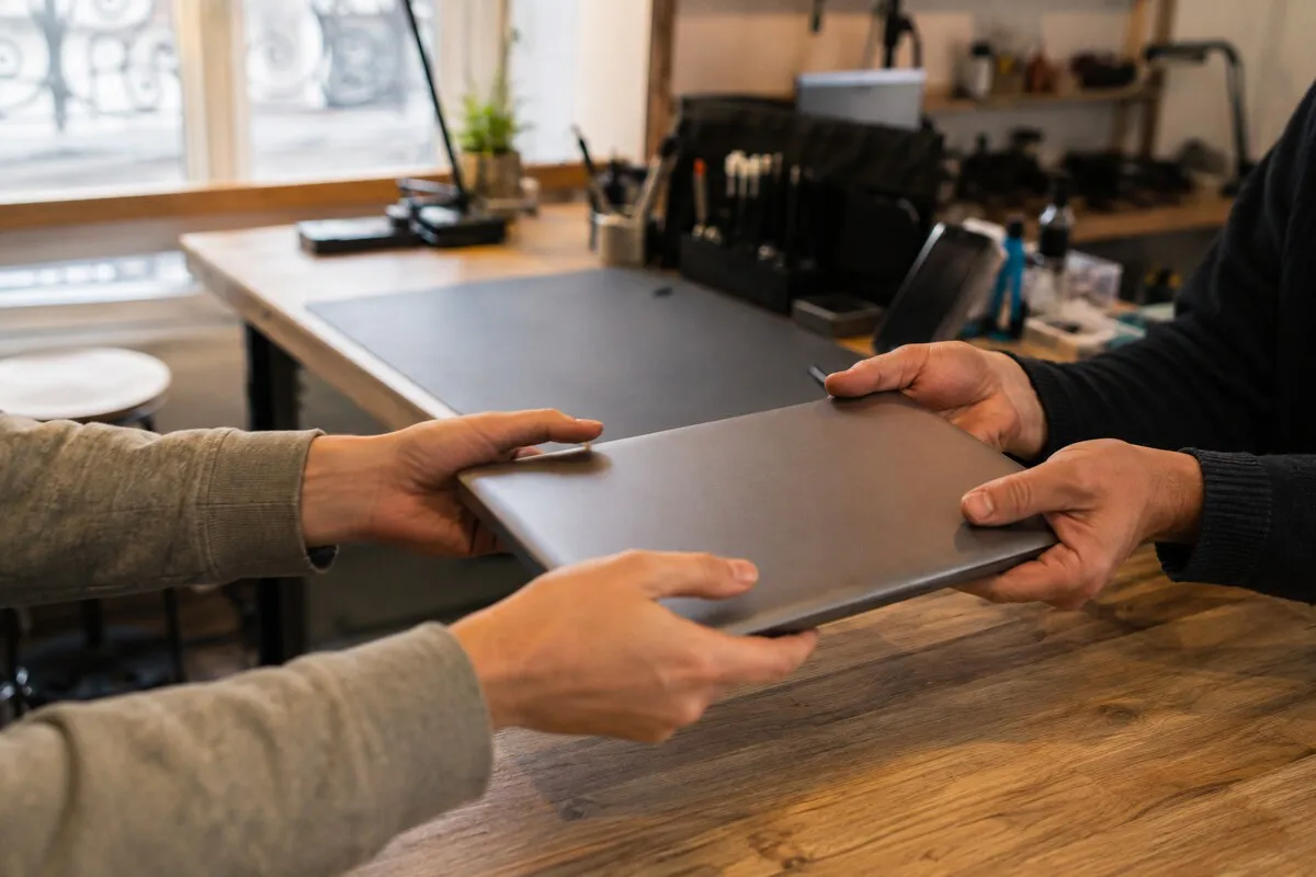 Two people passing a closed laptop across a counter, in a workshop space with tools in the background
