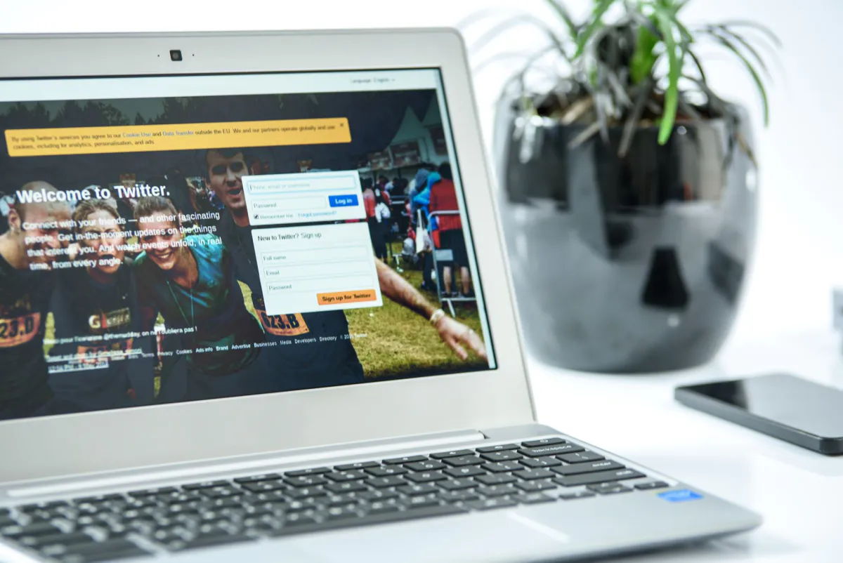 Open laptop showing a social media login page, with a potted plant and a phone resting beside it on a white desk