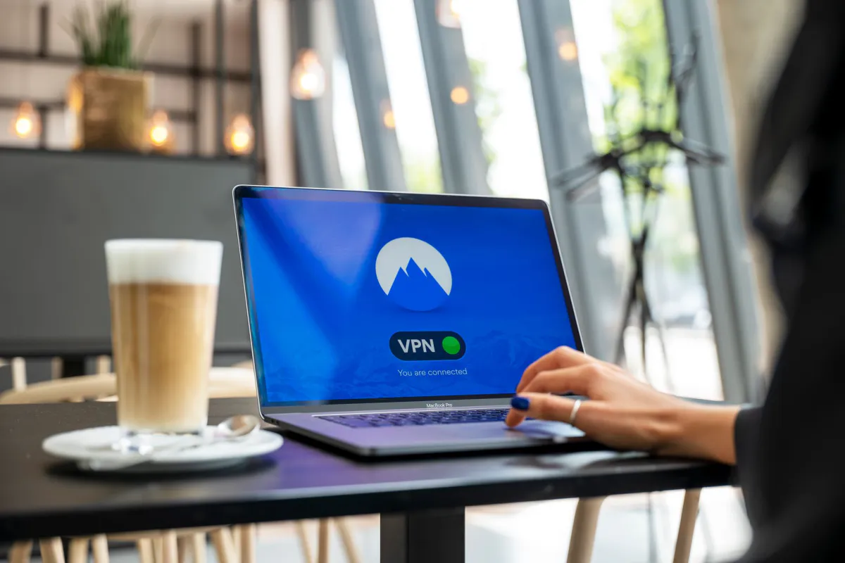 A hand on a laptop trackpad in a café, with a connected VPN app shown on screen and a coffee sitting on the table
