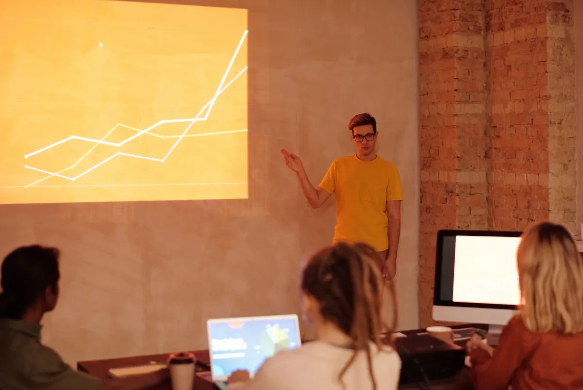 A man in a yellow t-shirt presenting to a small group, with a chart projected on a brick wall