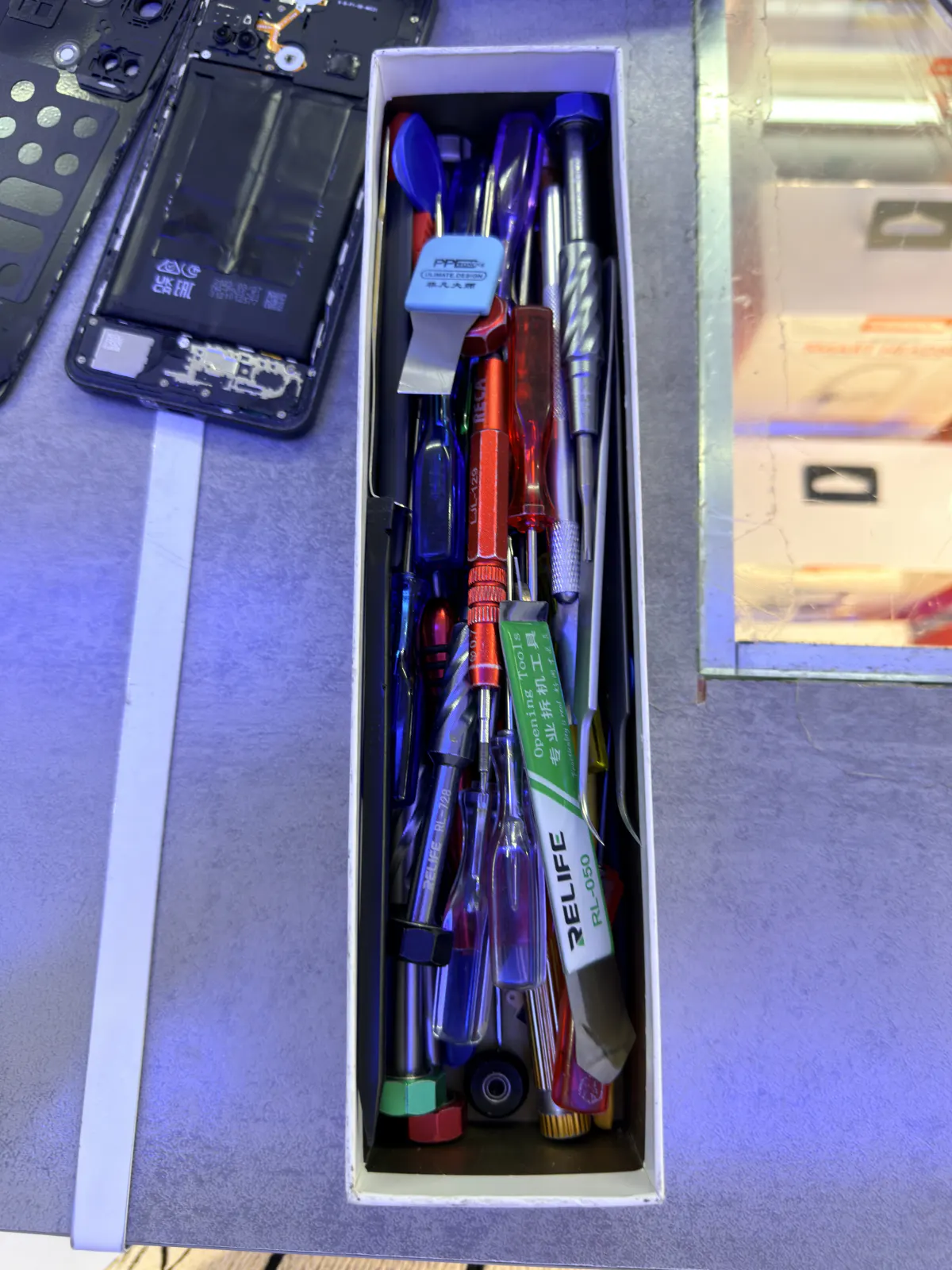 Open RELIFE storage box with precision screwdrivers, spudgers and a hex bit, next to a disassembled smartphone