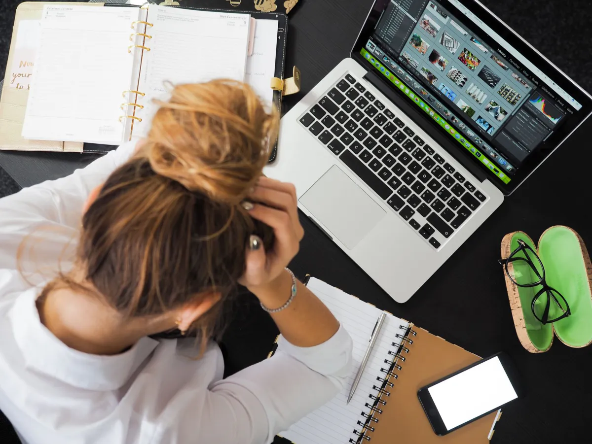 Woman sitting at a desk, head down with hands in her hair in front of an open laptop, surrounded by a planner and notebooks