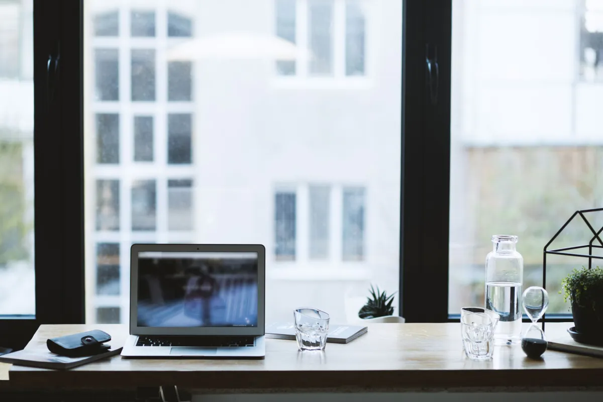 Ordinateur portable ouvert sur un bureau devant une grande fenêtre, avec verre d'eau et sablier posés à côté
