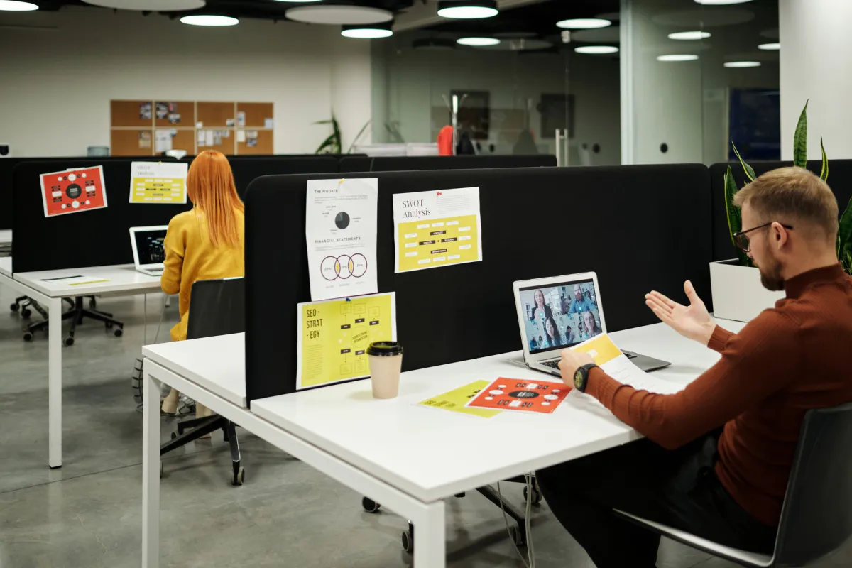 Open space avec deux personnes travaillant sur portable, l'une en visioconférence avec des collègues à distance, panneaux de notes affichés sur cloisons